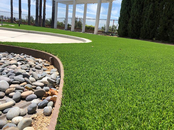 Close-up of neatly trimmed Artificial Grass of Casas Adobes beside a curved border of smooth gray river rocks in a landscaped outdoor area, with trees, pavement, and a white pergola visible in the background.