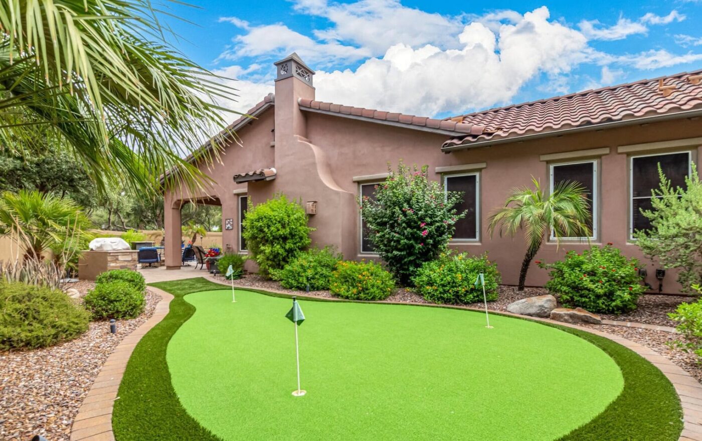 A backyard with Backyard Putting Greens, flagsticks, and landscaped plants beside a stucco house with a tile roof under a blue sky with clouds.