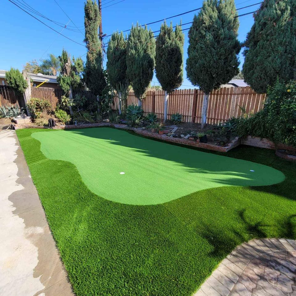 A backyard putting green with bright artificial turf is surrounded by trees, plants, and a wooden fence. Ideal for golf enthusiasts, these backyard putting greens feature two holes and are bordered by a concrete patio for added style.
