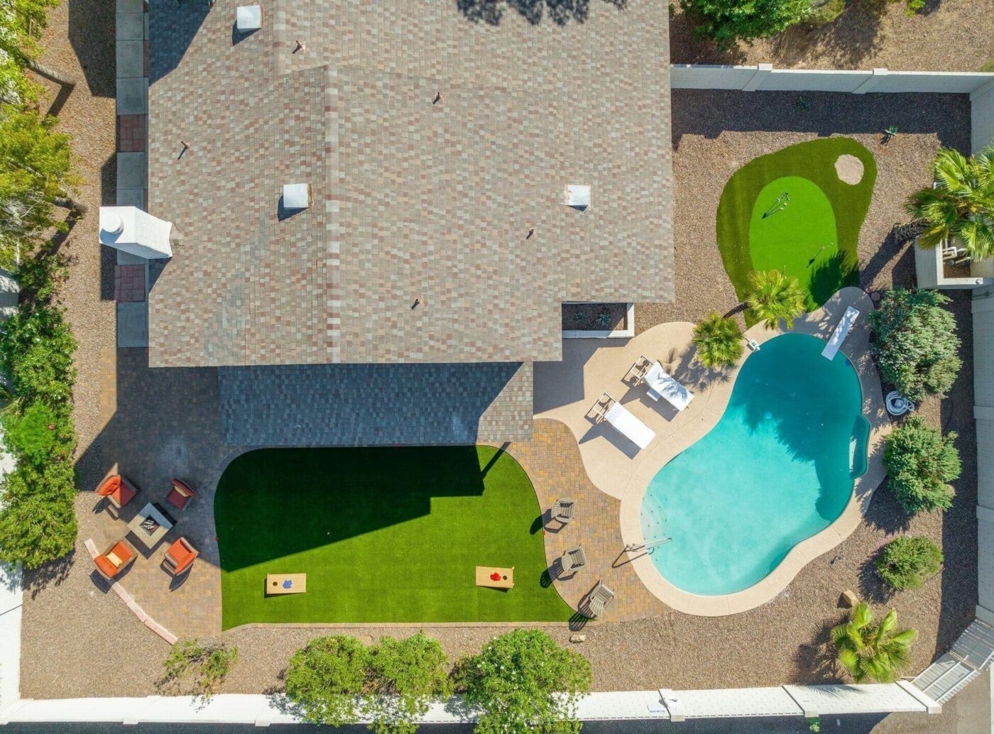 Aerial view of a backyard with a house, swimming pool, Backyard Putting Greens, artificial turf, cornhole boards, patio seating, and lush landscaping featuring bushes and trees.