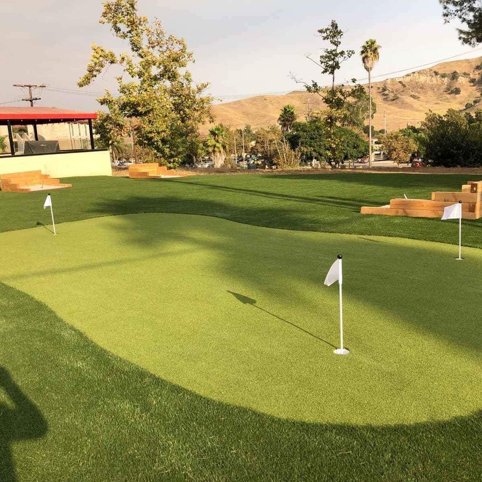 A small Backyard Putting Greens setup with three white golf flags on artificial turf, surrounded by well-maintained grass, wooden benches, trees, and hills in the background under a partly cloudy sky.