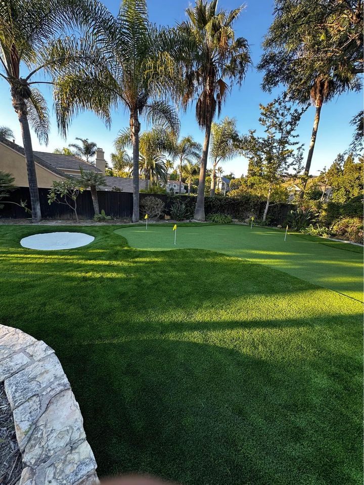 A lush Backyard Putting Green with several golf holes and yellow flags, bordered by palm trees and landscaping under a clear blue sky. A small sand bunker is visible on the left, creating a perfect oasis for golf enthusiasts.