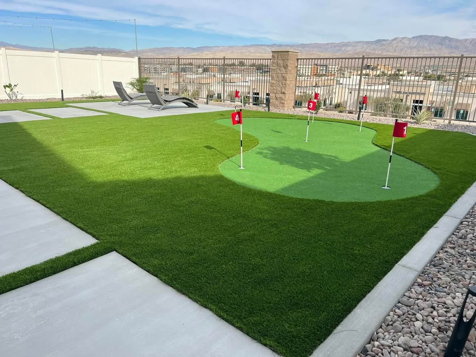 A backyard with artificial grass features Backyard Putting Greens with four red flags, lounge chairs on a concrete patio, and a view of desert mountains and houses beyond a metal fence.