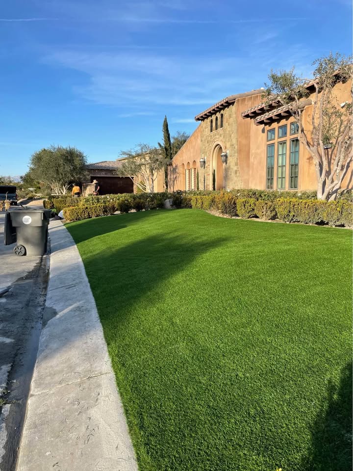 A large house with tan stucco walls and tall arched windows sits behind a neatly trimmed green lawn—Contact Artificial Grass of Casas Adobes for a lush, low-maintenance yard—and garbage bins are placed near the curb.