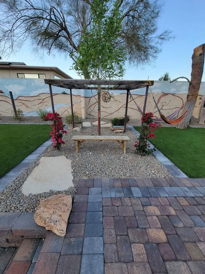 A backyard garden with a paved patio, a wooden bench under a pergola adorned with pink flowers, gravel ground, Artificial Grass of Casas Adobes, a hammock, and a mural-decorated wall in the background.
