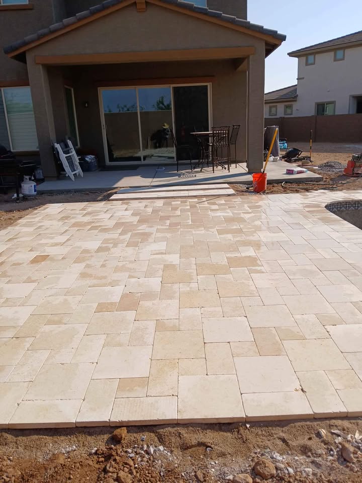 A newly installed light-colored stone patio, created through expert pavers installation, extends from the back of a house with sliding glass doors. Construction materials, tools, and dirt surround the patio, indicating ongoing landscaping work.