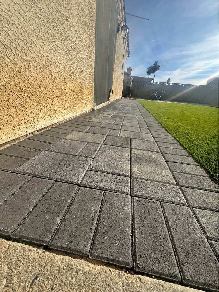 A paved walkway made with expert pavers installation features rectangular gray pavers running alongside a textured tan wall, adjacent to a bright green artificial grass lawn under a clear, sunny sky.