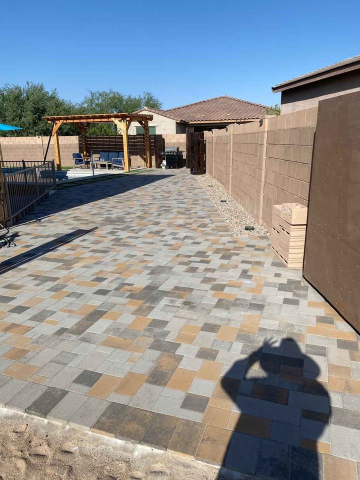 A backyard with a patterned paver patio from a recent pavers installation, a wooden pergola with seating, a block wall, and some stacked pavers. A person’s shadow is visible in the foreground beneath the clear blue sky.