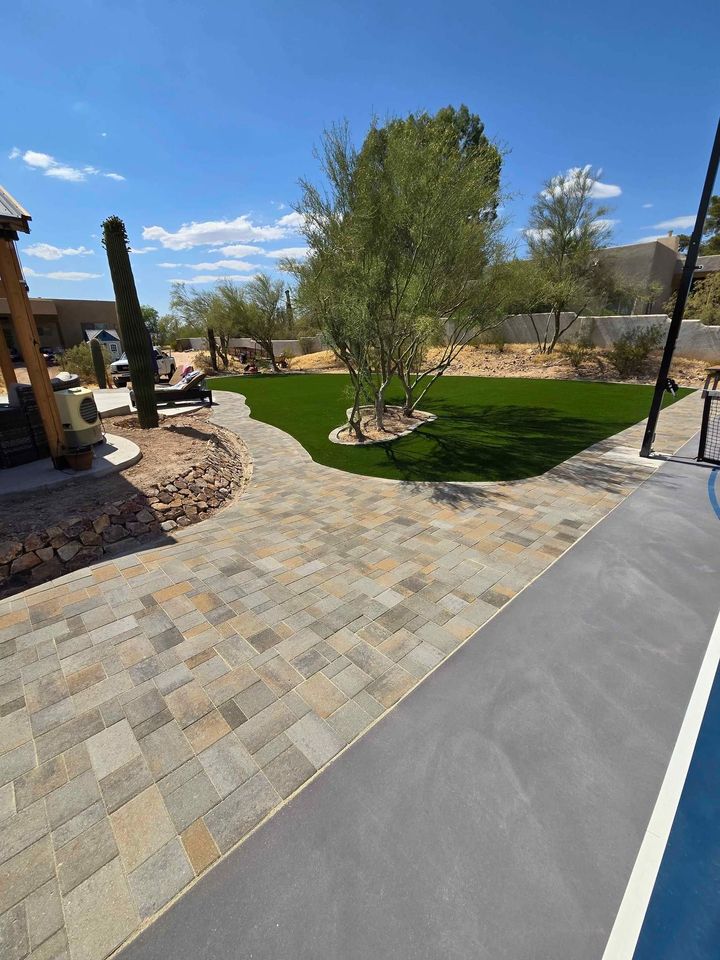 A backyard with expertly done pavers installation creating a stone walkway, artificial grass lawn, small desert trees, and a cactus under a clear blue sky. Landscaping rocks and features complete the inviting outdoor space.