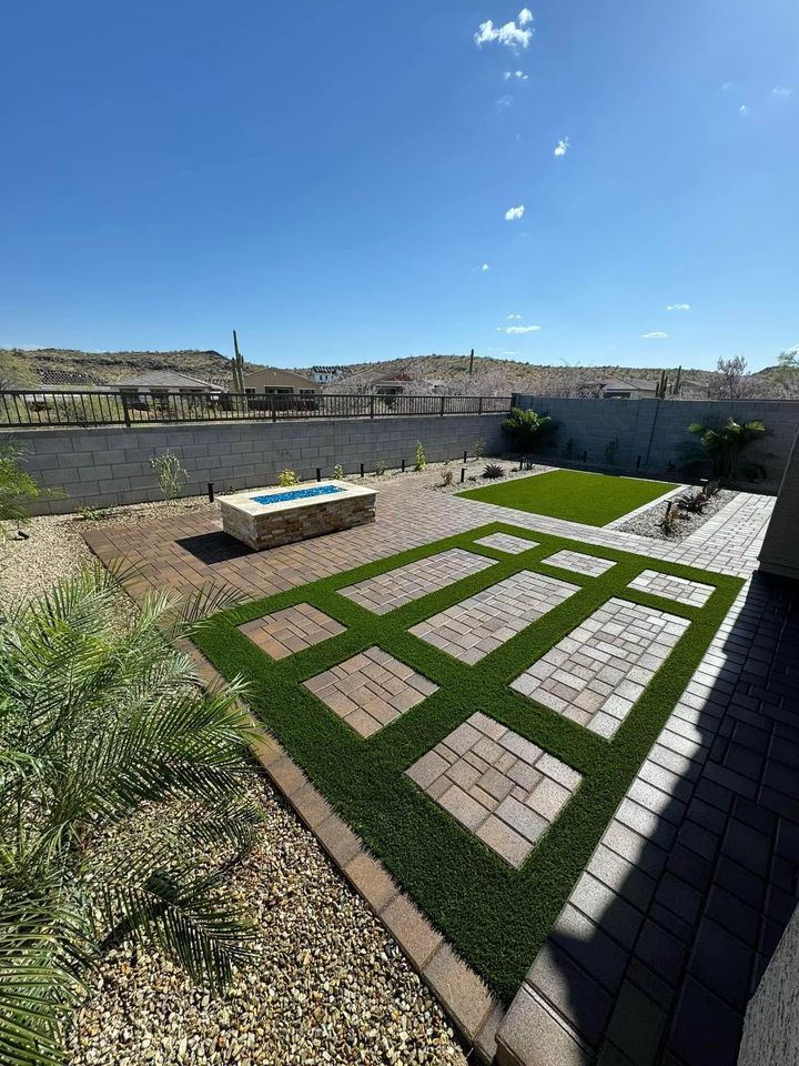 A backyard patio featuring expert pavers installation with geometric patterns of artificial turf and pavers, a rectangular firepit, desert landscaping, and views of a block wall with mountains and clear blue sky in the background.