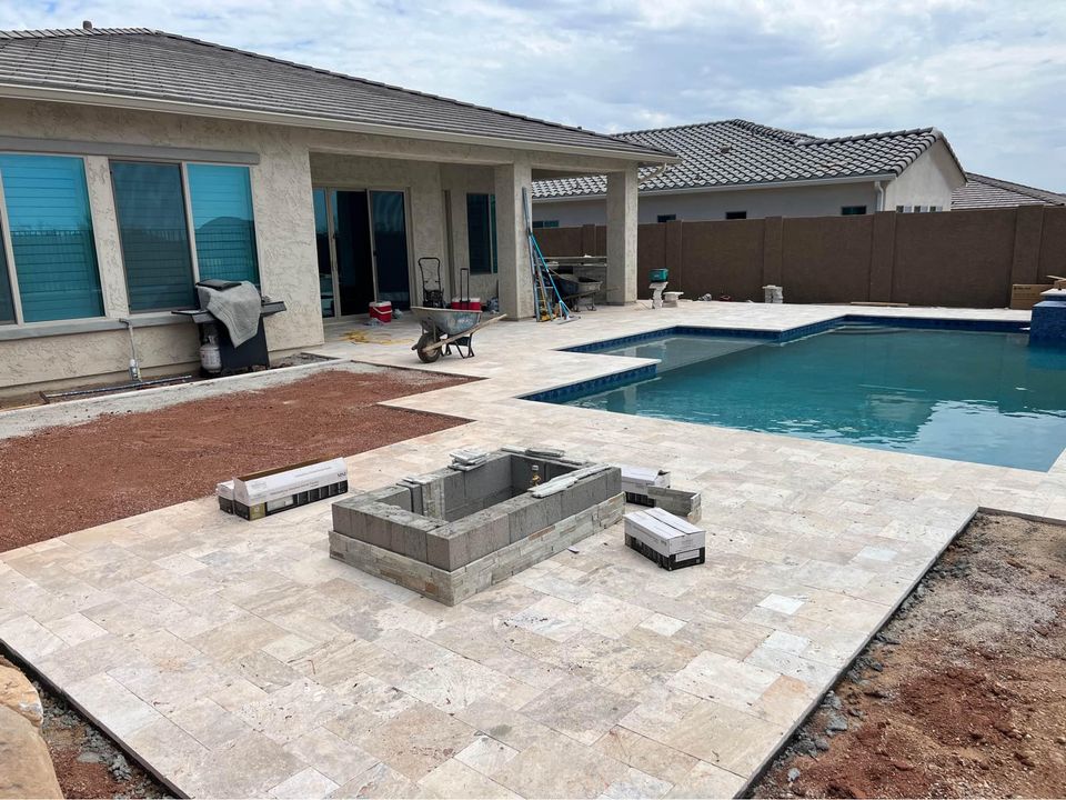 Backyard under construction with a partially built stone fire pit, tiled patio, ongoing pavers installation, swimming pool, and various construction materials near a beige house with large windows and a covered patio.