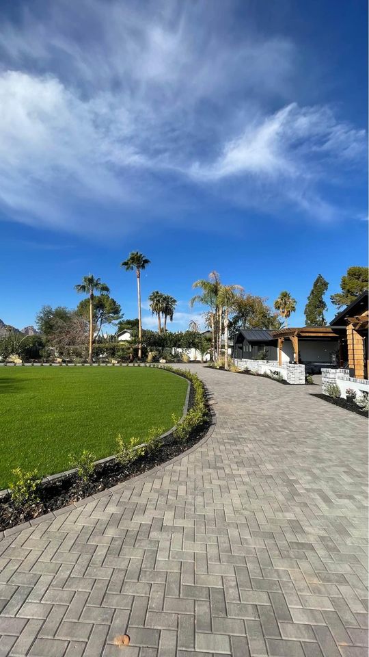 A wide brick pathway, showcasing expert pavers installation, curves beside a green lawn with palm trees and shrubs under a blue sky with wispy clouds; buildings are visible on the right side of the image.