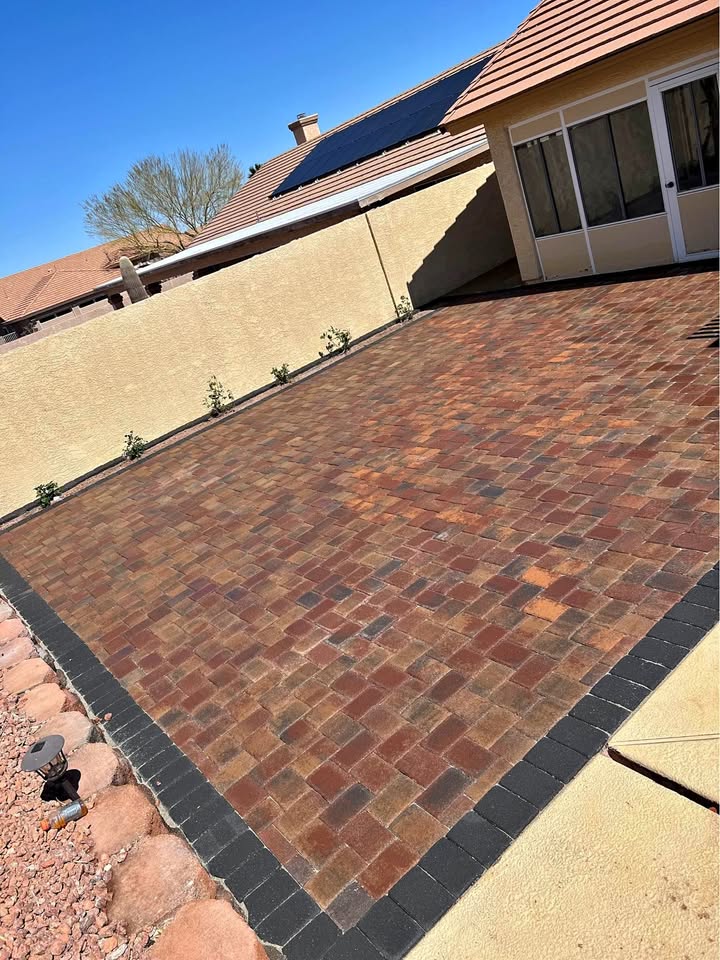 A backyard with a newly installed reddish-brown brick paver patio, featuring expert pavers installation bordered by dark bricks, sits next to a beige house with a screened-in porch and tan stucco wall. Small plants line the wall; rocky landscaping is on the left.