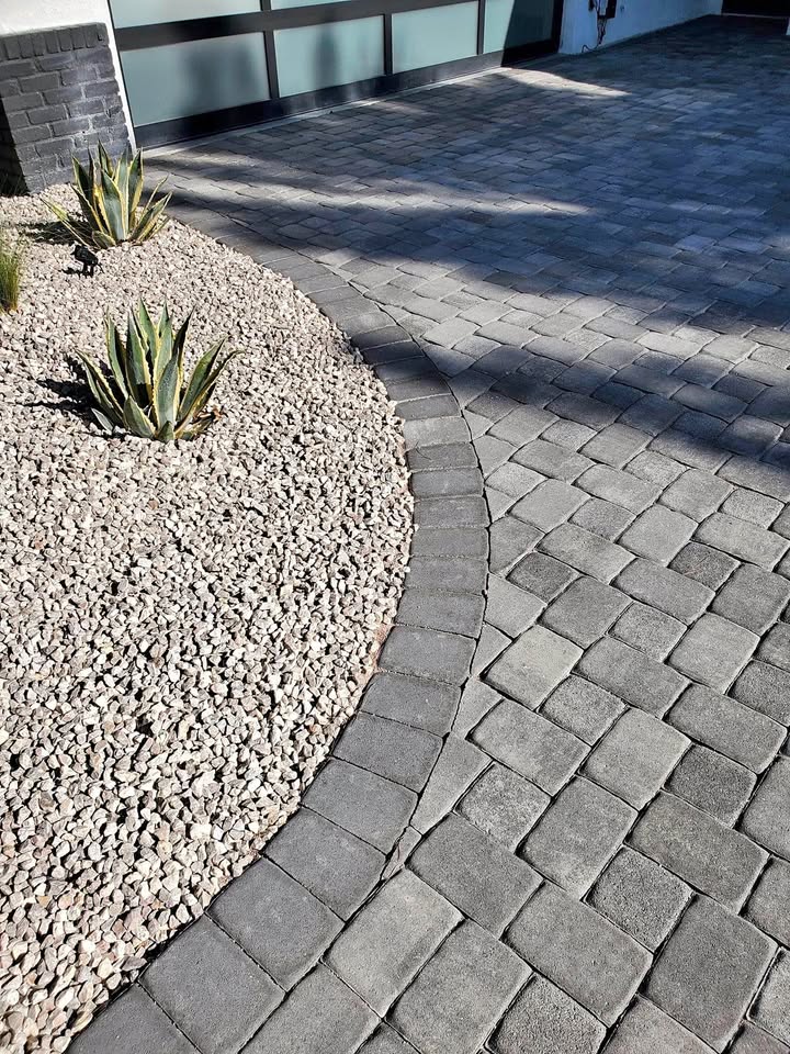A curved edge separates a cobblestone-patterned driveway, created with expert pavers installation, from a landscaped area with light-colored gravel and two agave plants near a modern building.