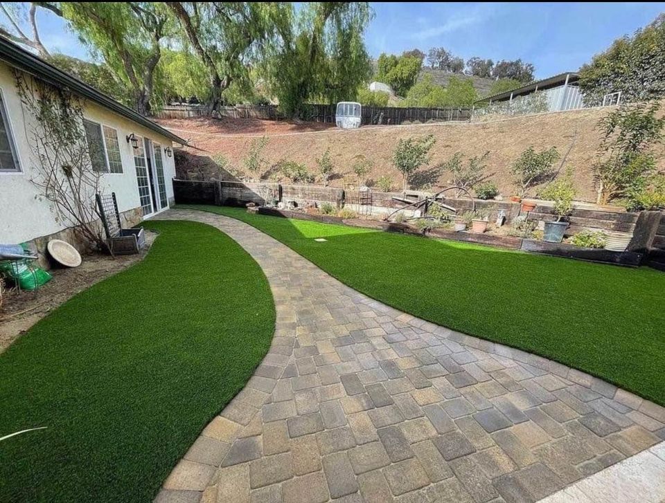 A backyard featuring a curved stone pavers installation divides two sections of artificial green grass, bordered by garden beds, potted plants, and a sloped hillside with trees and shrubs in the background.