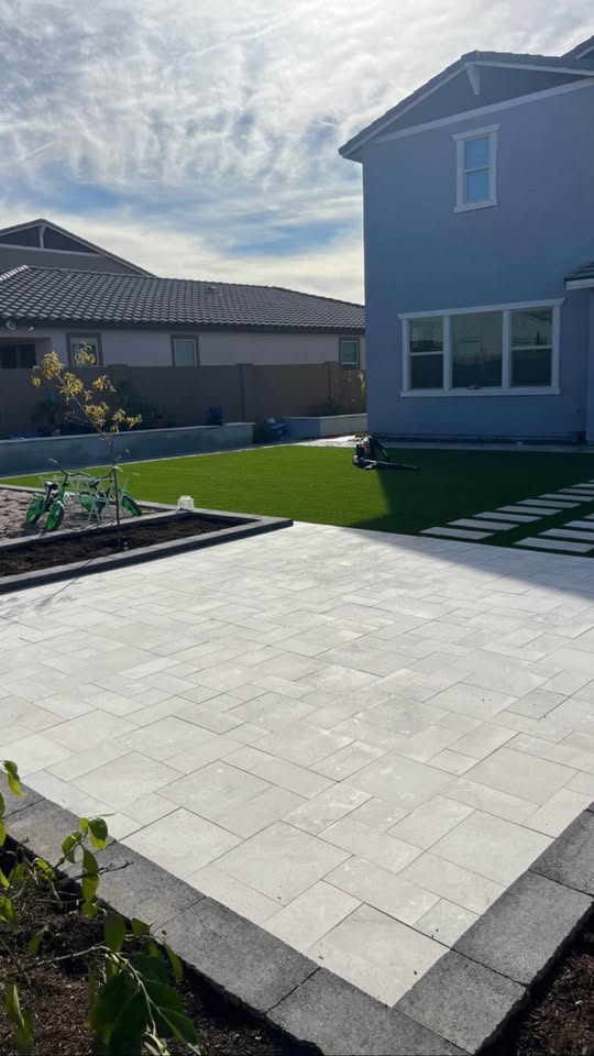 A backyard with professional pavers installation featuring light-colored stone pavers, artificial grass, a gray two-story house, a bicycle, and a swing set under a partly cloudy sky.