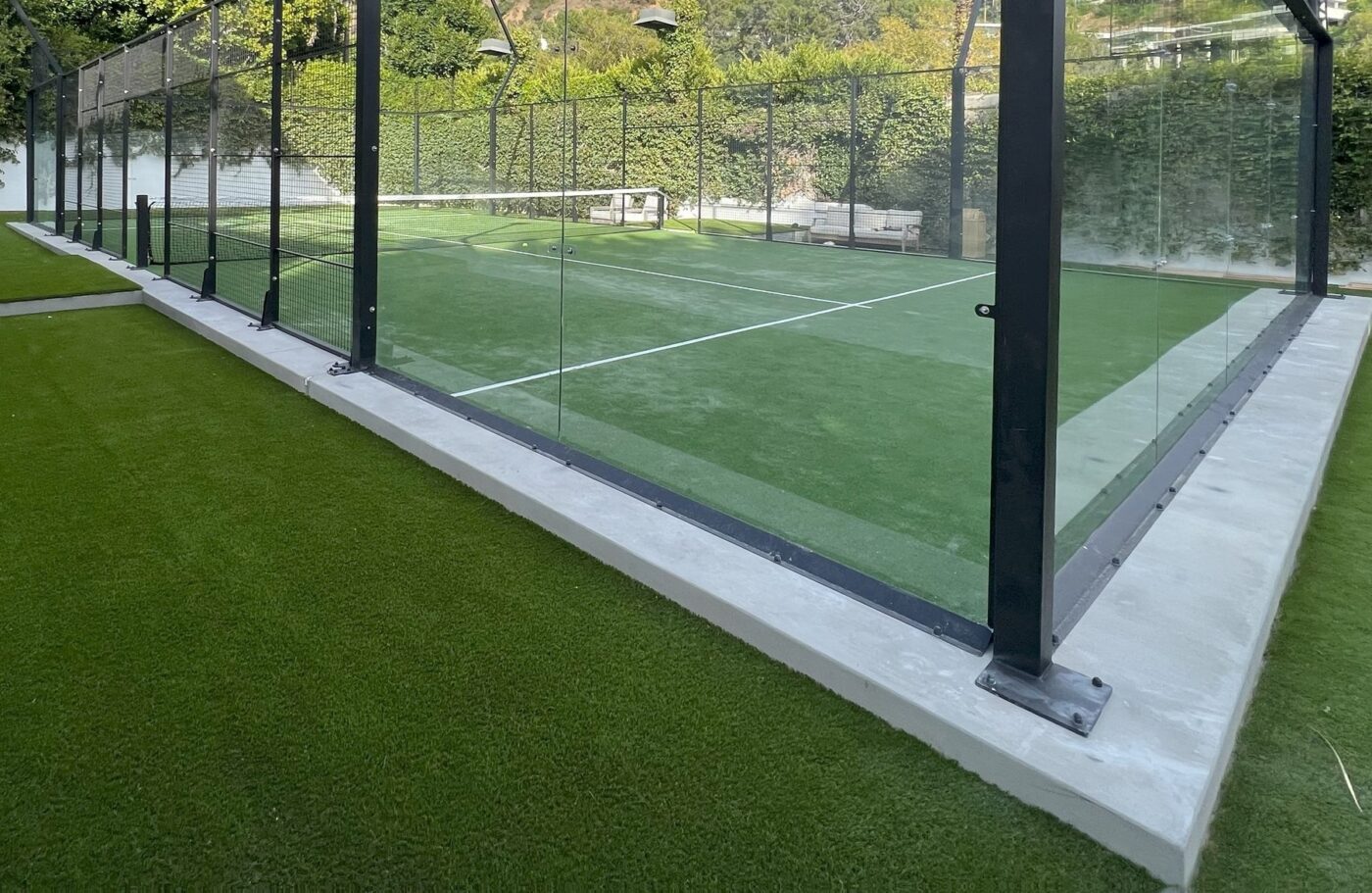 A modern padel court with glass walls and sports field artificial turf, surrounded by greenery and trees, viewed from an outside angle.