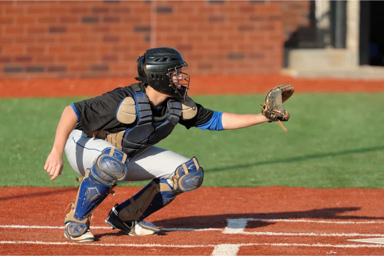 A baseball catcher in full gear squats behind home plate, wearing a helmet and mitt, ready to catch a pitch on a sports field with artificial turf and a brick wall in the background.