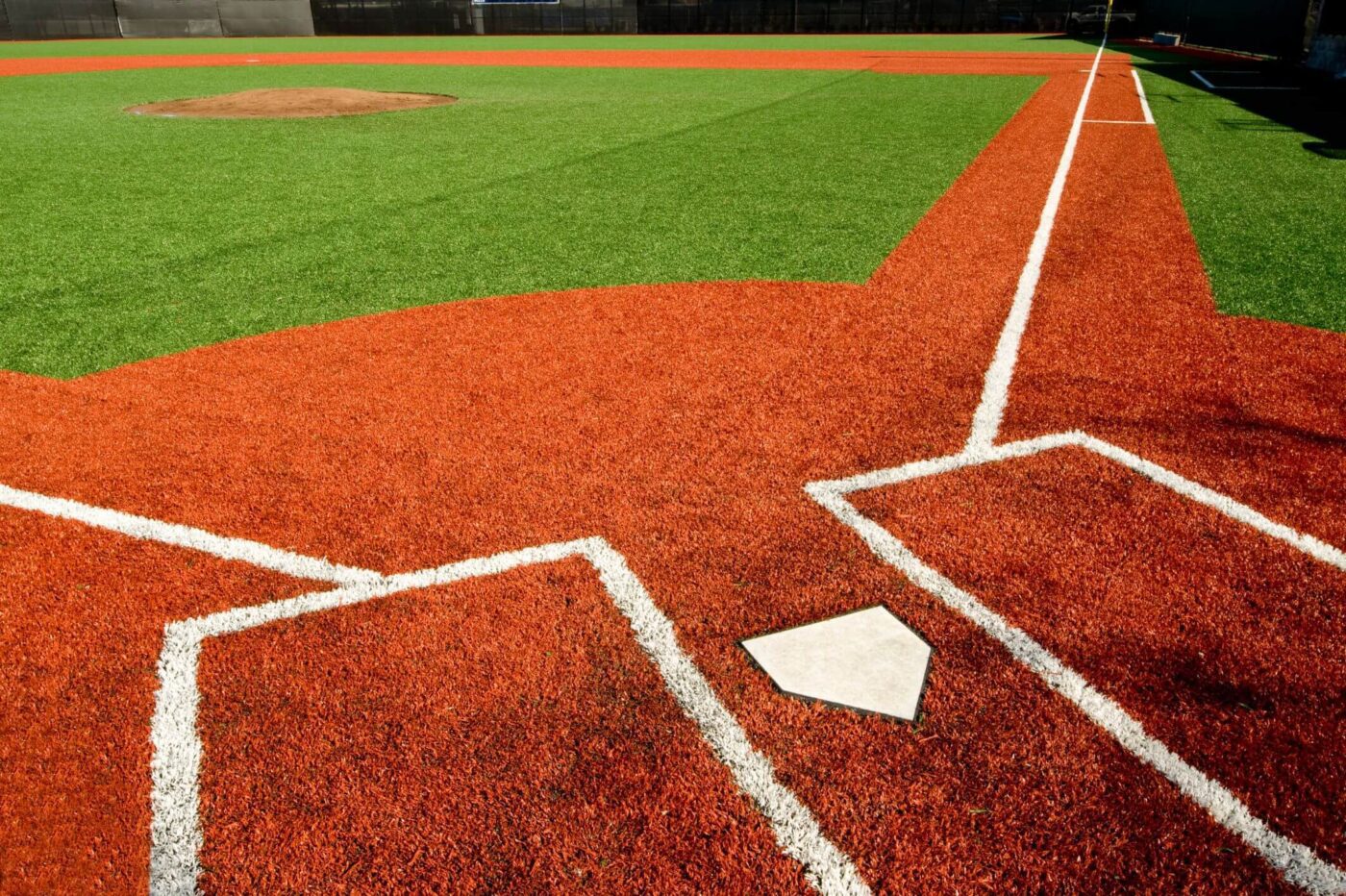 A close-up view of a sports field with bright green artificial turf, focusing on home plate, batter's boxes, and the foul lines extending toward the pitcher's mound in the distance.