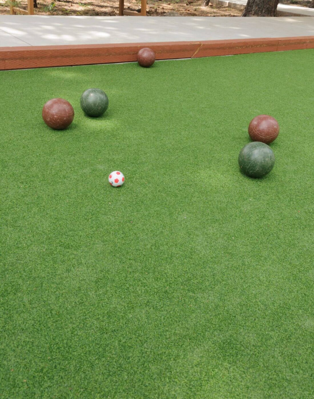 Four bocce balls and a small white-and-red pallino ball rest on the artificial turf sports field, framed by wood borders and trees in the background.
