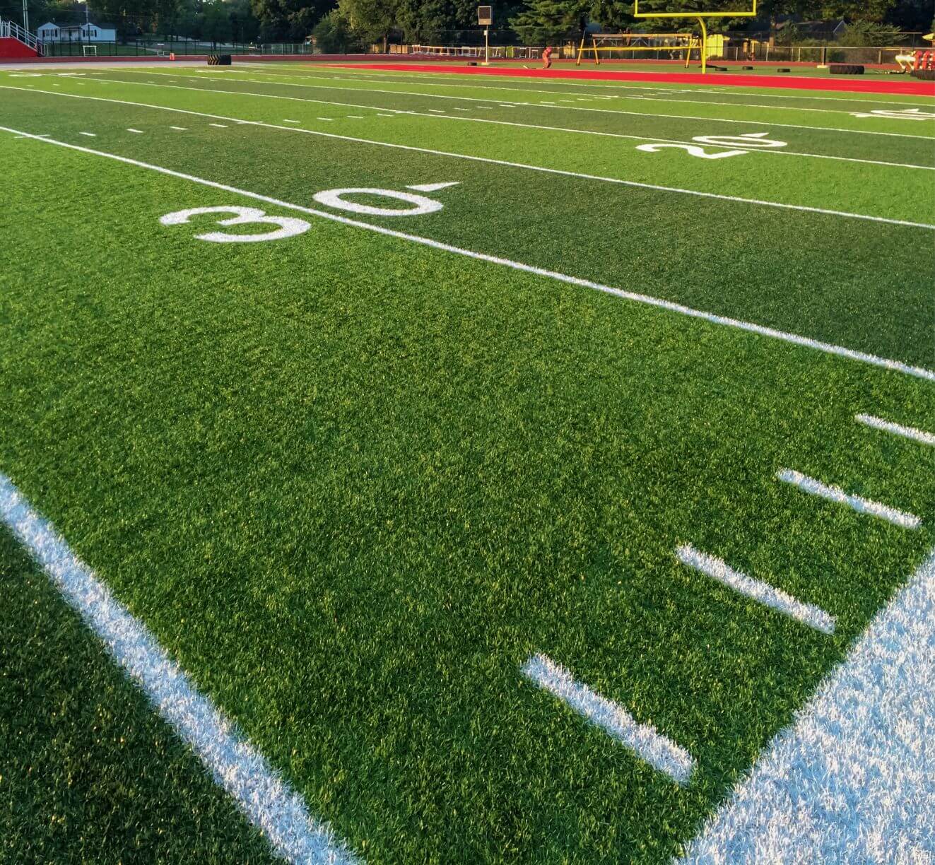 A close-up view of a sports field features yard lines and numbers on green artificial turf under sunlight, with a yellow goalpost and red track visible in the background.